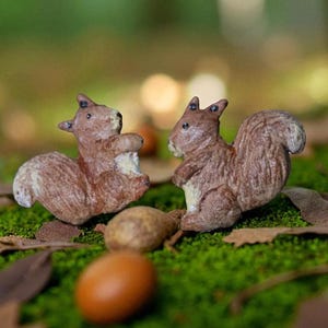 May include: Two brown squirrel figurines with fluffy tails are positioned on a bed of green moss, surrounded by fallen leaves and acorns. The squirrels face each other, creating a charming woodland scene. The figurines are detailed and textured.