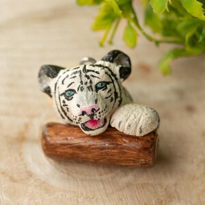 Peut inclure: Une figurine de tigre blanc, un ourson avec des yeux bleus, reposant sa tête sur une bûche brune. La figurine est en pâte polymère et est une création artisanale.