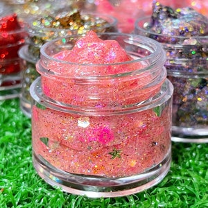 May include: Close-up of a pink lip gloss with glitter in a clear jar. The lip gloss is surrounded by other jars of lip gloss in different colors.