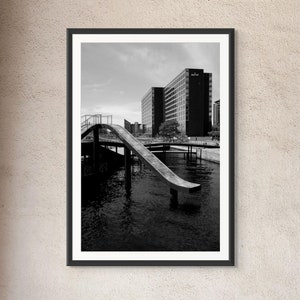 May include: A black and white photograph of a modern building with a long, curved ramp leading down to a body of water. The ramp is made of concrete and has a metal railing. The building is tall and has many windows. The water is calm and reflects the sky.