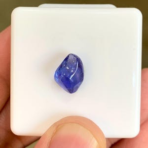 以下が含まれることがあります： 小さく、カットされた、濃い青色の宝石が白い正方形の上に置かれています。 宝石は研磨され、反射する表面を持っています。 石は手で持たれており、白い正方形がコントラストのある背景を提供しています。