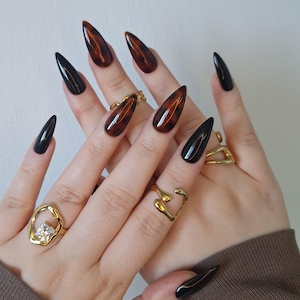 May include: Close-up of hands displaying long, pointed artificial nails. Some nails are black, others feature a tortoiseshell design. Gold rings are worn on the fingers, and a gold chain bracelet is visible. The nails are a fashion accessory.