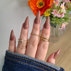 Könnte beinhalten: Eine Hand mit langen, mandelförmigen, braunen Nägeln. Die Finger sind mit goldenen Ringen geschmückt, darunter ein Band aus kleinen Perlen, ein Doppelband mit einem rautenförmigen Akzent und ein Kreuzmuster. Die Hand trägt eine Jeansjacke, und Blumen sind im Hintergrund.