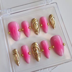 Pode incluir: Um conjunto de dez unhas postiças rosa e douradas com efeito degradê. As unhas têm um detalhe dourado na metade superior da unha e uma pequena pérola dourada na metade inferior.