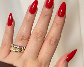 以下が含まれることがあります： アーモンド型の爪に光沢のある鮮やかな赤色のマニキュアが塗られた手。薬指には2つの指輪が重ね付けされており、小さな透明な石がセットされた細いシルバーのバンドと、花の形をした透明な石のクラスターがあしらわれた幅広のゴールドのバンドが特徴です。