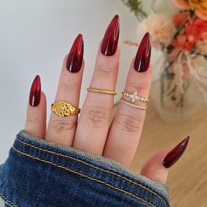 Könnte beinhalten: Eine Hand mit langen, stilettoförmigen, burgunderfarbenen Ombre-Nägeln. Die Hand ist mit drei goldenen Ringen geschmückt: einem strukturierten Band, einem zarten Ring mit Kreuzdesign und einem detaillierten Siegelring. Die Person trägt eine Jeansjacke.