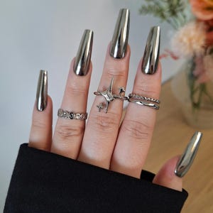Puede incluir: Una mano con uñas largas de cromo plateado. Las uñas son largas y puntiagudas, y el acabado cromado les da un aspecto brillante y metálico. La mano lleva varios anillos plateados.