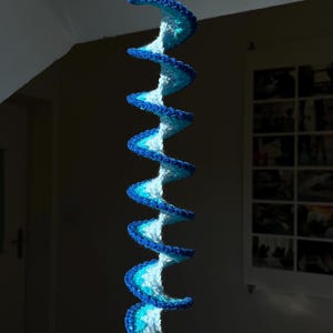 Peut inclure: Décoration suspendue en spirale, réalisée en crochet. La spirale présente un dégradé de bleus et de blanc, avec une bordure bleu foncé. Une perle transparente en forme de goutte d'eau est suspendue en bas. Décoration intérieure.