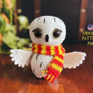 Può includere: Gufo bianco all'uncinetto con grandi occhi neri che indossa una sciarpa a righe rosse e gialle. Schema all'uncinetto PDF.