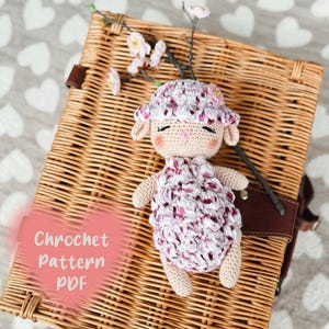 Può includere: Schema a uncinetto PDF per un amigurumi di agnello in crochet rosa e bianco. L'agnello è sdraiato su un cesto di vimini con un gambo floreale dietro.