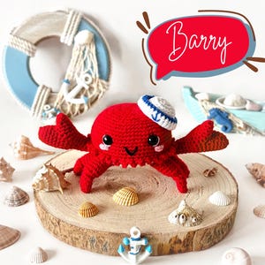 Può includere: Un granchio rosso all'uncinetto che indossa un cappello da marinaio blu e bianco. Il granchio è seduto su una fetta di legno con conchiglie intorno. Il testo "Barry" è scritto in un fumetto sopra il granchio.
