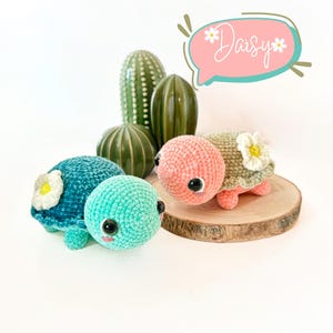 Può includere: Due peluche di tartaruga all'uncinetto con occhi ricamati e dettagli floreali. Le tartarughe sono turchese e rosa e sono sedute su una fetta di legno. La parola "Daisy" è scritta in un fumetto sopra le tartarughe.