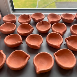 Könnte beinhalten: Eine Sammlung von Terrakotta-Diya-Lampen, traditionellen indischen Öllampen, die auf einer Oberfläche angeordnet sind. Die Diyas haben eine leuchtend orange-rote Farbe und eine abgerundete Form mit einem kleinen Ausguss für den Docht.