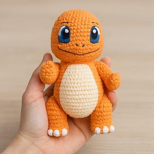 Könnte beinhalten: Ein handgehäkeltes Charmander-Spielzeug in Orange mit blauen Augen und einem cremefarbenen Bauch. Die Plüschfigur hat weiße Akzente an den Füßen und wird in einer Hand gehalten. Das Spielzeug ist ein beliebter Charakter aus dem Pokémon-Franchise.