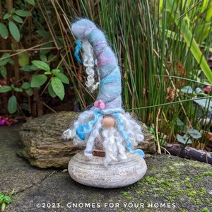 Könnte beinhalten: Eine skurrile Gnomfigur mit einem blauen und rosafarbenen Wirbelhut, weißem Bart und blauen lockigen Haaren. Der Gnom sitzt auf einem grauen Stein.
