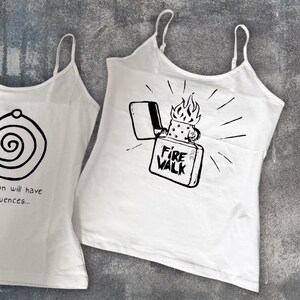 May include: Two white tank tops with thin straps. One features a black spiral design, and the other displays a black graphic of a lighter with flames and the text "FIRE WALK". The tops are made of a lightweight fabric.