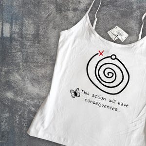 May include: White camisole with thin straps featuring a black spiral design with a red X and the text "This action will have consequences...". A small butterfly graphic is also present. A tag is attached.