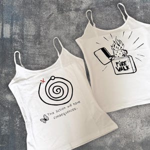 May include: Two white tank tops with black graphic designs. One features a spiral with the text "This action will have consequences..." and a butterfly. The other has a lighter with the text "Fire Walk".