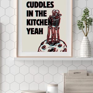 May include: A framed print with a white background and black text that reads "Remember cuddles in the kitchen yeah". The print features a black and white illustration of two skeletons hugging on a moon-like surface.