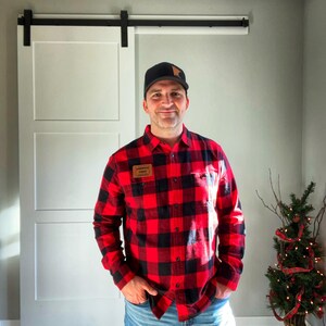 Könnte beinhalten: Ein Mann trägt ein rot-schwarzes kariertes Flanellhemd mit einem braunen Lederflicken, auf dem "American Made" auf der linken Brust steht. Er trägt auch eine schwarze Baseballkappe mit einem braunen Aufnäher auf der Vorderseite. Er steht vor einer weißen Scheunentür.