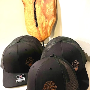 Peut inclure: Trois casquettes de camionneur noires et vertes en mesh avec un logo brun qui dit "Pizza Factory" sur le devant. Les casquettes sont sur une surface blanche.