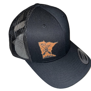 Op de afbeelding: Zwarte truckercap met een bruine leren patch met een omtrek van de staat Minnesota met de woorden "USA MN" en een paar gekruiste peddels.
