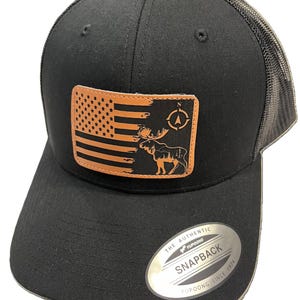 May include: Black and brown trucker hat with a leather patch featuring an American flag, a moose, and a compass. The hat has a mesh back and a "The Authentic Yupoong Snapback Yupoong Since 1974" label.