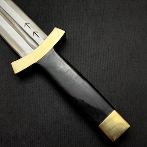 May include: A close-up of a dagger with a polished steel blade and a black handle. The handle is accented with gold-colored metal at the guard and pommel. Two arrows are etched on the blade.