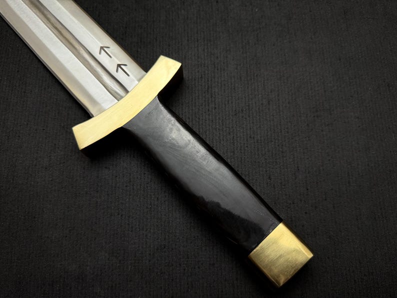 May include: A close-up of a dagger with a polished steel blade and a black handle. The handle is accented with gold-colored metal at the guard and pommel. Two arrows are etched on the blade.