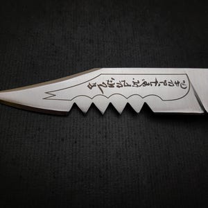 May include: A silver knife with a serrated edge and a black handle. The blade has an inscription in Arabic script that reads "وَأَنْتَ عَلَىٰ خَيْرٍ كَثِيرٍ".