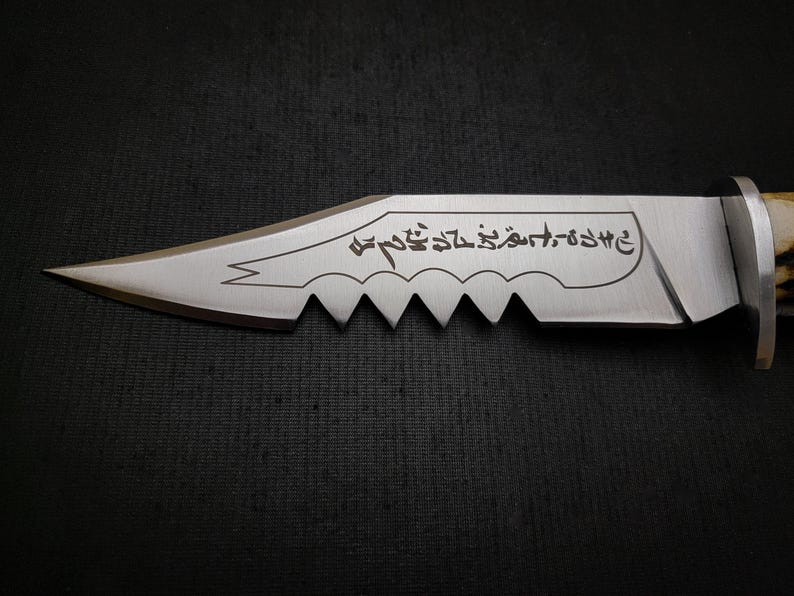 May include: A silver knife with a serrated edge and a black handle. The blade has an inscription in Arabic script that reads "وَأَنْتَ عَلَىٰ خَيْرٍ كَثِيرٍ".