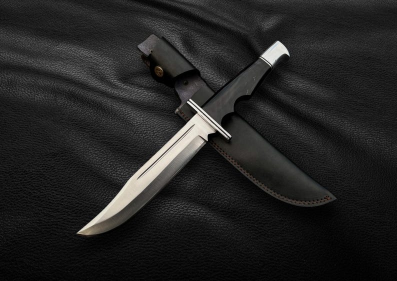 May include: A silver hunting knife with a black leather sheath. The knife is lying on a black leather surface.