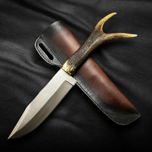 May include: A hunting knife with a brown leather sheath and a handle made of antler. The knife has a silver blade and a gold accent on the handle.