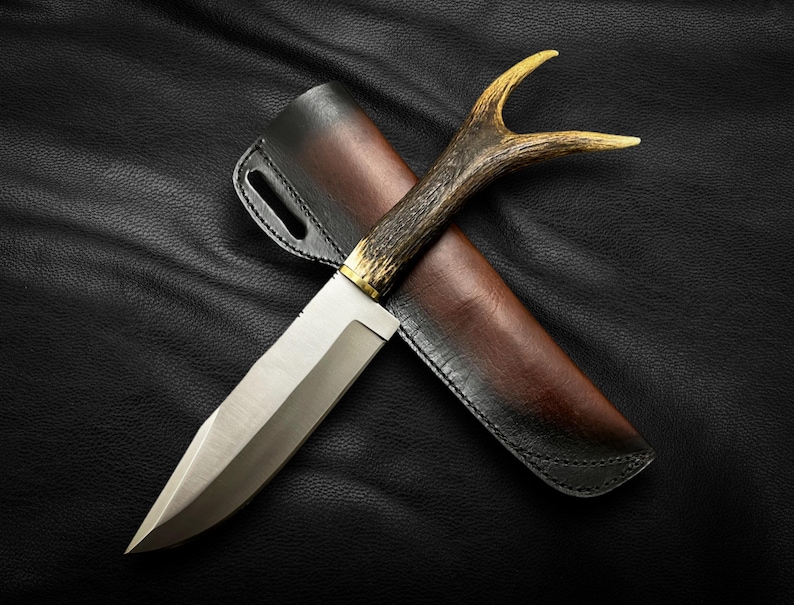 May include: A hunting knife with a brown leather sheath and a handle made of antler. The knife has a silver blade and a gold accent on the handle.