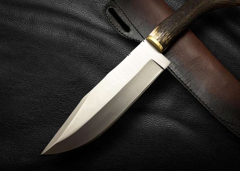 May include: A silver-colored hunting knife with a brown antler handle and a leather sheath. The knife is lying on a black leather surface.