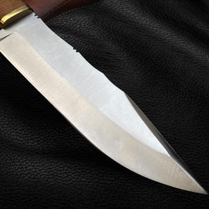 May include: A close-up of a silver knife with a wooden handle. The knife is sharp and has a serrated edge. The knife is lying on a black leather surface.