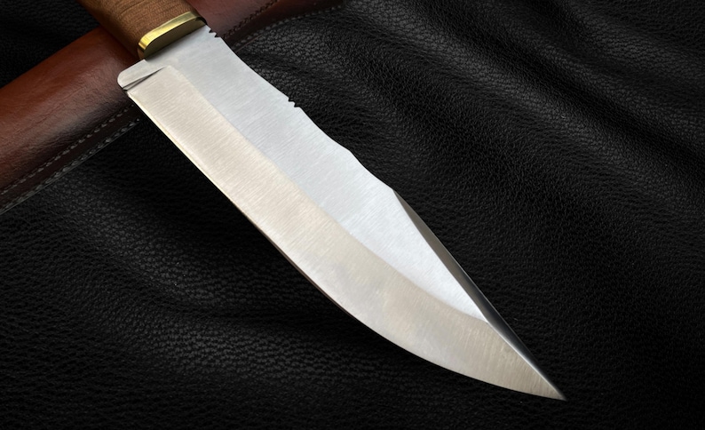 May include: A close-up of a silver knife with a wooden handle. The knife is sharp and has a serrated edge. The knife is lying on a black leather surface.
