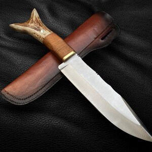 May include: A hunting knife with a brown leather sheath and a stag antler handle. The blade is silver and has a serrated edge.