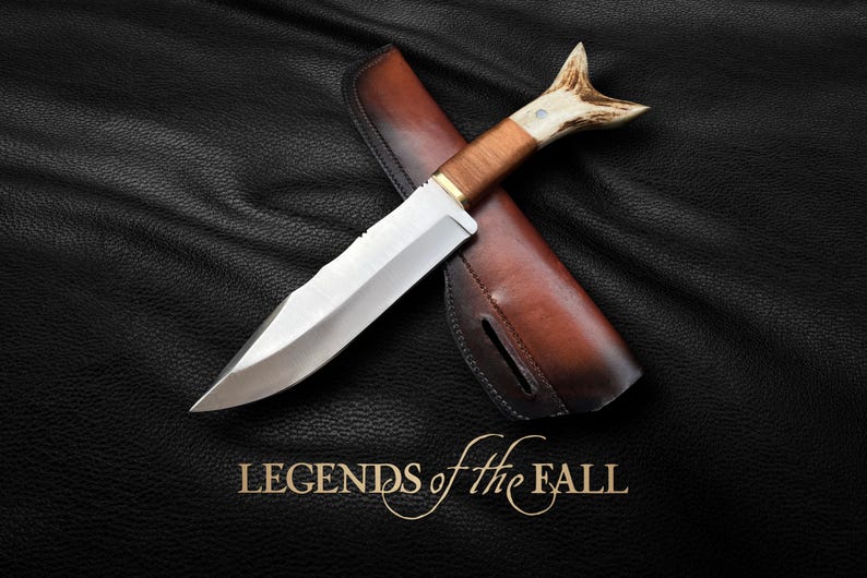 May include: A hunting knife with a brown leather sheath and a stag antler handle. The knife has a silver blade and the text "LEGENDS of the FALL" is printed below the knife.