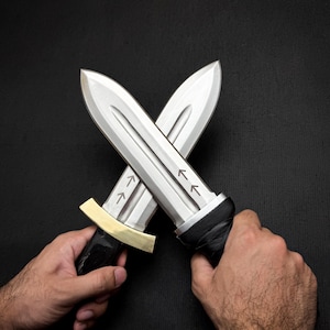 May include: Two crossed silver daggers with black handles and gold accents. The blades have arrow markings. The daggers are held against a black background.