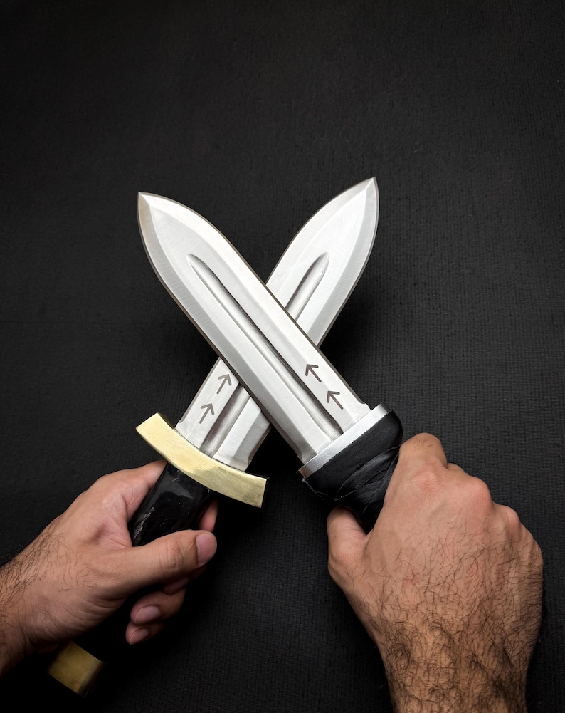 May include: Two crossed silver daggers with black handles and gold accents. The blades have arrow markings. The daggers are held against a black background.