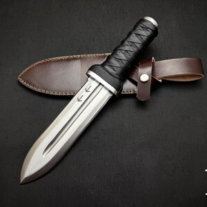 May include: A silver dagger with a double-edged blade and a black leather-wrapped handle rests on a brown leather sheath. The dagger has two arrows etched on the blade. The sheath has a brown leather belt loop.