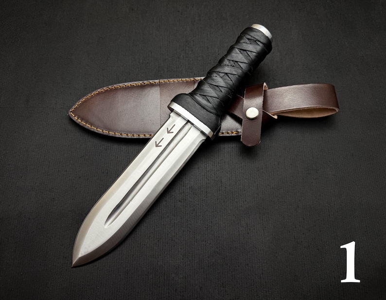May include: A silver dagger with a double-edged blade and a black leather-wrapped handle rests on a brown leather sheath. The dagger has two arrows etched on the blade. The sheath has a brown leather belt loop.