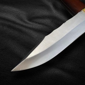 May include: A close-up of a silver knife blade with a wooden handle. The knife is lying on a black leather surface.