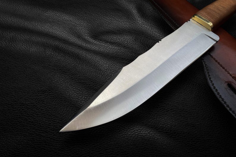 May include: A close-up of a silver knife blade with a wooden handle. The knife is lying on a black leather surface.
