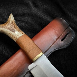 May include: A brown leather sheath with a stitched edge holds a hunting knife with a brown wooden handle and a stag antler pommel.