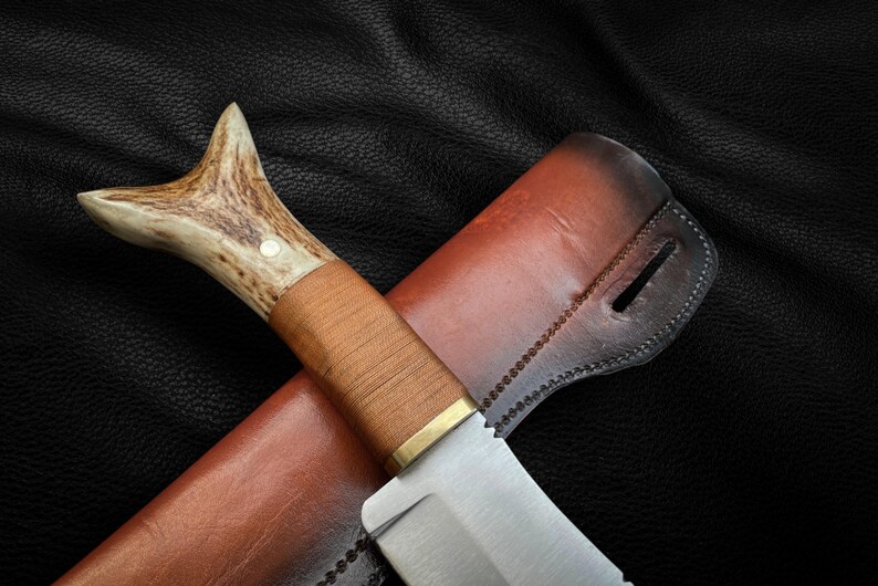 May include: A brown leather sheath with a stitched edge holds a hunting knife with a brown wooden handle and a stag antler pommel.