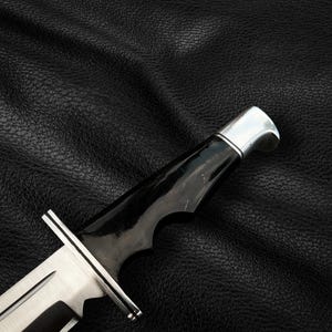 May include: A black handled hunting knife with a silver blade and a black leather background.