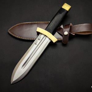 May include: A dagger with a silver blade, black handle, and gold accents, resting on a brown leather sheath. The dagger has two arrows etched on the blade. The sheath has a brown leather belt loop.