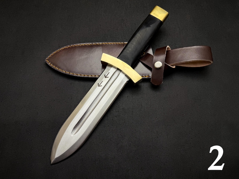 May include: A dagger with a silver blade, black handle, and gold accents, resting on a brown leather sheath. The dagger has two arrows etched on the blade. The sheath has a brown leather belt loop.